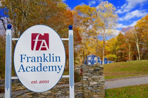 The entrance sign at Franklin Academy, with the school's logo, an F and A inside a red square, and the words Franklin Academy in blue. In the background are the yellows and oranges of New England fall foliage.