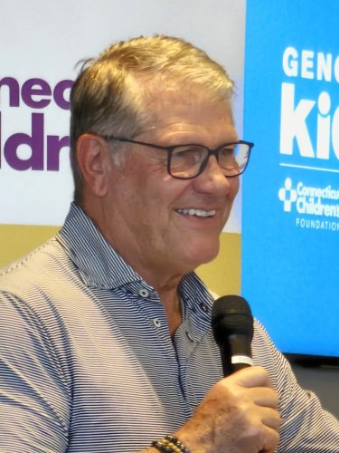 UConn women's basketball coach Geno Auriemma, a white man wearing black glasses and a plain gray button-down shirt, holding a microphone. In the background are the two logos of Connecticut Children's Medical Hospital and Geno For The Kids.