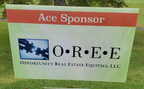 The puzzle pieces logo of OREE, Opportunity Real Estate Equities LLC, on the light green sign of a Watkinson Golf Classic 2024 Ace Sponsor.