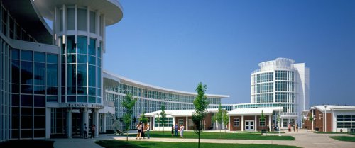 The campus of Connecticut State Community College (previously known as Manchester Community College) on a clear, blue day. The photo is taken from a 3/4 angle, with elegant, sleek, white, many-windowed buildings on the left side. The landscape is dotted with trees.